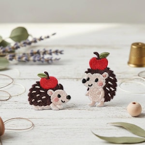 May include: Two embroidered hedgehog patches. Each hedgehog has a brown spiky back, a tan face with pink cheeks, and a red apple on its head. The patches are on a white surface with other decorative items.