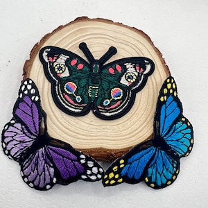 May include: Three embroidered butterfly patches in vibrant colors. One is dark green with pink and yellow accents, the others are purple and blue with black outlines. The patches are displayed on a wooden slice.