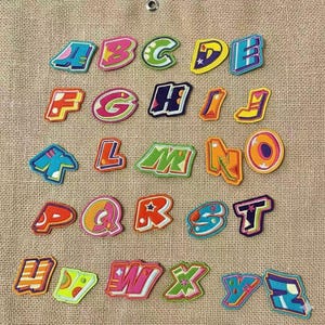 May include: A collection of colorful, embroidered alphabet letters in various designs. Each letter is outlined in a contrasting color and features a unique pattern, with a mix of blues, greens, pinks, and yellows. The letters are arranged on a textured, tan fabric.