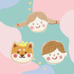 Op de afbeelding: Geborduurde patches met een meisje met vlechten, een Shiba Inu hond met een kroon en een jongen met bruin haar. Elke patch heeft een lachend gezicht en roze wangen. De patches staan op een pastelkleurige achtergrond.