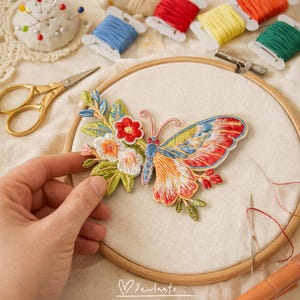 Puede incluir: Una colorida mariposa bordada con detalles florales se muestra en un bastidor de bordado. La mariposa tiene alas rojas, naranjas, azules y amarillas. Hilos y tijeras doradas en el fondo.