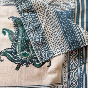 May include: A beige textile with a large teal paisley design and teal geometric borders. The fabric has a soft, natural appearance, with a detailed floral pattern in the center. The borders feature intricate geometric and floral motifs.