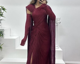 May include: A burgundy pleated gown with a high slit and long sleeves. The dress features a draped bodice and a flowing skirt. The sleeves extend into cape-like panels. The dress is floor-length and appears to be made of a lightweight fabric.
