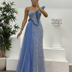 Puede incluir: Vestido de noche largo, color azul claro. El corpiño está adornado con lentejuelas y una superposición de tul fluido añade volumen a la falda. Una correa decorativa se extiende sobre un hombro, creando una silueta elegante. El vestido mide unos 150 cm de largo.