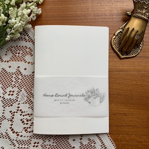 May include: A white journal wrapped with a band featuring the text "Home Bound Journals" and botanical illustrations. The journal measures 5 x 7 inches (13 x 18 cm) and contains 60 pages. The journal is on a wooden surface with a decorative hand-shaped object.