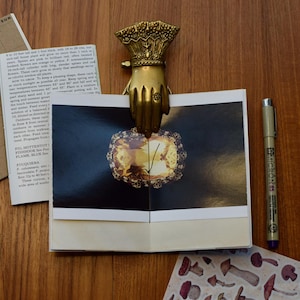 May include: A closed book with a gold metal decorative clasp in the shape of a hand. The book has a yellow and brown image on the front cover. The book is sitting on a wooden surface.
