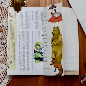 May include: An open book with a colorful illustration of a bear holding an umbrella with a bird and a red bird on top. The text on the page includes the words "wren, fox, loon, coon, pony, dog, frog, hare, bear, dowpane."