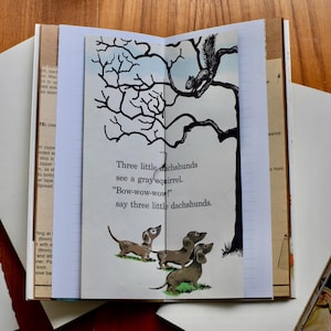 May include: An open book with a page showing a black and white illustration of three dachshunds and a squirrel in a tree. The text on the page reads "Three little dachshunds see a gray squirrel "Bow-wow-wow!" say three little dachshunds."