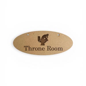 Könnte beinhalten: Ovales Holzschild mit den Worten "Throne Room" in einer Serifenschrift. Eine Silhouette einer Person, die auf einer Toilette sitzt, befindet sich über dem Text. Das Schild hat zwei kleine Löcher oben zum Aufhängen.