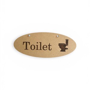 Könnte beinhalten: Ovales Holzschild mit dem Wort "Toilet" in dunkelbraunen Buchstaben und einer Toilettengrafik. Das Schild hat zwei kleine Löcher oben zum Aufhängen. Der Hintergrund ist weiß.