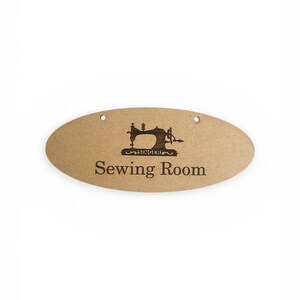 Könnte beinhalten: Ovales, hellbraunes Holzschild mit den Worten "Sewing Room" in dunkelbrauner Schrift. Eine Silhouette einer Vintage Singer Nähmaschine befindet sich über dem Text. Das Schild hat drei kleine Löcher oben zum Aufhängen.