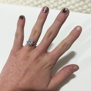 May include: A silver ring with a floral design is worn on a hand with painted nails. The ring features a detailed, openwork pattern. The nails are painted with a dark, metallic polish. The hand is resting on a white surface.