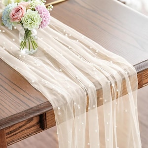 May include: A sheer, cream-colored table runner adorned with scattered faux pearls. The runner is draped over a dark wooden table, with a small glass vase of pastel flowers in the upper left corner. The runner adds a touch of elegance to any table setting.