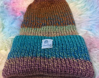 May include: A handmade knit beanie in shades of brown, teal, and purple. The hat features a ribbed brim and a tag that reads "Handmade". The beanie is displayed on a fluffy, multicolored surface.