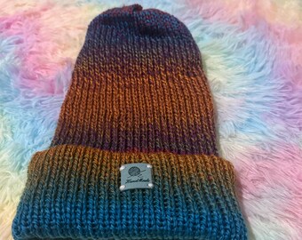 May include: A hand-knitted beanie hat with a color gradient from blue to orange to purple. The hat has a folded brim and a small tag that reads "Hand Made". The hat is made of a soft, textured yarn.