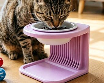 Puede incluir: Un soporte elevado de color violeta claro para cuenco de gato con un cuenco de cerámica blanca que contiene comida seca. El soporte cuenta con un soporte vertical acanalado y una base plana. Tres pequeñas bolas de fieltro texturizadas en verde, rojo y azul se encuentran cerca.