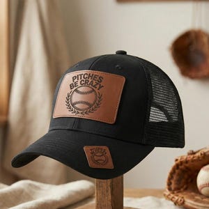Könnte beinhalten: Schwarze Baseballkappe mit Mesh-Rückseite und Lederaufnäher. Der Aufnäher zeigt den Text "Pitches Be Crazy" über einem Baseball und Lorbeerkranz-Design. Ein kleinerer Aufnäher mit Baseball-Design befindet sich auf der Vorderseite.