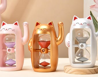 May include: Three cat-shaped hourglasses in pink, gold, and white. Each cat has a raised paw and holds an hourglass with colored sand. The pink hourglass has purple sand, the gold has red sand, and the white has gray sand.