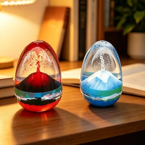 May include: Two egg-shaped glass ornaments, one red and one blue, are displayed on a wooden surface. The red piece has a volcano design, the blue one a mountain. Both contain liquid and glitter, creating a visual effect.
