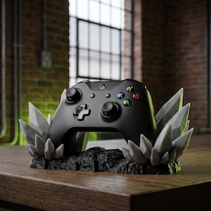 Puede incluir: Mando Xbox negro sobre un soporte decorativo. El soporte tiene una base negra con formaciones de cristal gris. El mando tiene botones de colores y una cruceta. El soporte está sobre una mesa de madera.