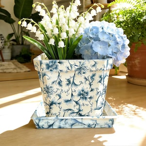 Puede incluir: Una maceta cuadrada de cerámica con un diseño floral azul y blanco, que contiene flores blancas de lirios de los valles y hortensias azules. La maceta se encuentra sobre un platillo a juego. Otras plantas están en el fondo.