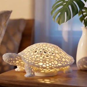 Może przedstawiać: Biała lampa w kształcie żółwia z ażurowym wzorem skorupy. Lampa jest podświetlana od wewnątrz, dając ciepłą poświatę. Stoi na drewnianym stoliku nocnym, obok wazonu z zielonymi liśćmi.