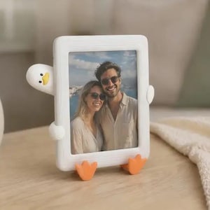 May include: A white picture frame with a duck design, holding a photo of a couple. The frame has a duck head on the side, orange feet at the bottom, and a yellow beak. The frame is sitting on a wooden surface.