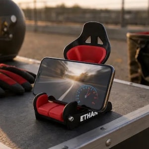 May include: A black and red smartphone stand shaped like a racing bucket seat. The stand features a small headrest and seat base with red cushioning. The word ETHAN is printed in white on the side of the black base, which holds a smartphone.