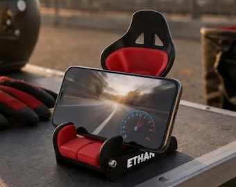Puede incluir: Un soporte para smartphone negro y rojo con forma de asiento de carreras. El soporte cuenta con un pequeño reposacabezas y una base de asiento con acolchado rojo. La palabra ETHAN está impresa en blanco en el lateral de la base negra.