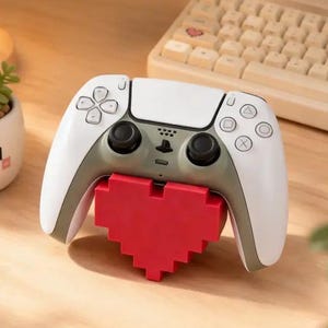Peut inclure: Une manette de jeu blanche et grise avec un support rouge en forme de cœur pixélisé. La manette comporte des boutons noirs et un logo PlayStation. Un clavier et une petite plante en pot sont également visibles.