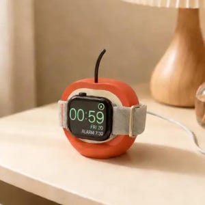 Può includere: Un supporto di ricarica a forma di mela rossa per smartwatch. Il supporto presenta uno stelo nero sulla parte superiore e un cinturino grigio chiaro avvolto attorno al centro. Il quadrante digitale mostra l'ora 00:59, Fri 20 e Alarm 7:30.