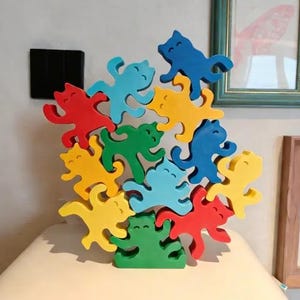 Op de afbeelding: Een kleurrijk houten kattenstapelspeelgoed. De katten zijn in verschillende kleuren, waaronder rood, geel, groen en blauw. Het speelgoed is gestapeld op een wit oppervlak. Een ingelijste foto hangt op de achtergrond.