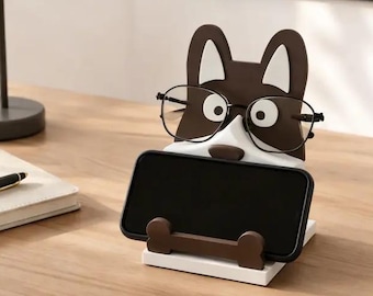 Puede incluir: Soporte para teléfono con forma de perro marrón y blanco con gafas. El soporte sujeta un smartphone negro y tiene un soporte en forma de hueso. El soporte está sobre una base blanca.