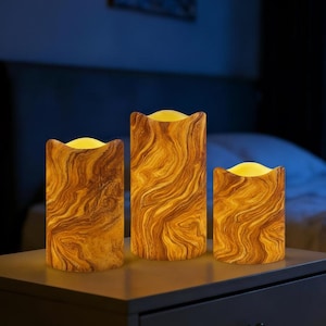 May include: Set of three battery-operated LED candles with a wood-grain design. The candles have a warm, yellow glow and are arranged on a nightstand. The tallest candle is approximately 6 inches tall.