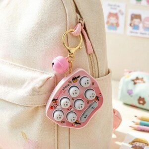 May include: A pink, pentagonal fidget toy keychain attached to a cream-colored canvas backpack. The toy features seven white, circular push-buttons with small faces, a gold-tone metal ring, and a pink bell. The text "Guess where I am" is printed.