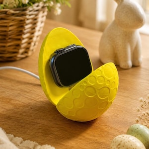 May include: A yellow, egg-shaped charging station with a black smartwatch inside. The station is open, revealing the watch's screen. A white charging cable extends from the station. Easter eggs and a decorative bunny are in the background.