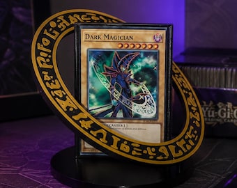 Puede incluir: Una carta coleccionable Dark Magician en una funda protectora, colocada en un soporte circular negro y dorado con símbolos místicos grabados. La carta muestra una figura con túnica y bastón sobre un fondo mágico oscuro con estrellas.