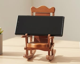 May include: A miniature wooden rocking chair phone stand with a medium brown finish. The chair features a slatted backrest, armrests, and curved rockers at the base, currently holding a black rectangular smartphone in a horizontal position.