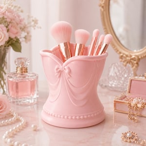 Puede incluir: Un soporte para brochas de maquillaje de color rosa claro con un diseño moldeado tipo corsé, detalles de tela drapeada y un pequeño lazo en el centro. El soporte contiene cinco brochas con virolas de oro rosa y cerdas rosas.