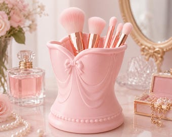 Puede incluir: Un soporte para brochas de maquillaje de color rosa claro con un diseño moldeado tipo corsé, detalles de tela drapeada y un pequeño lazo en el centro. El soporte contiene cinco brochas con virolas de oro rosa y cerdas rosas.