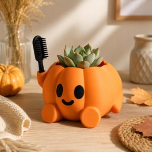 May include: A bright orange pumpkin-shaped planter with a smooth finish, featuring a smiling face with black oval eyes and a curved mouth. A small black plastic comb is inserted into a dedicated slot on the side, and a green succulent plant sits inside.