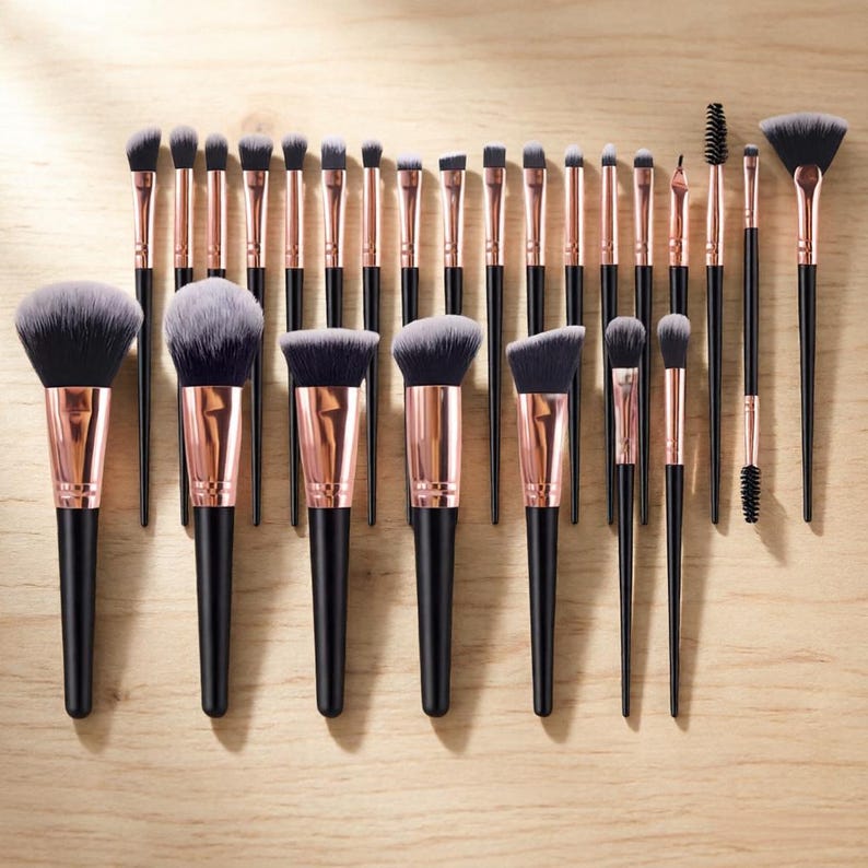 May include: A collection of makeup brushes with black handles and rose gold ferrules. The brushes vary in size and shape, including powder, blush, and eyeshadow brushes. The bristles are a mix of black and white, arranged on a light wood grain surface.