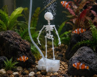 Puede incluir: Un adorno de acuario con forma de esqueleto blanco de plástico sobre una base rectangular, conectado a un tubo de aire transparente que libera burbujas desde el cráneo. La figura está rodeada de rocas volcánicas, plantas acuáticas y peces payaso.