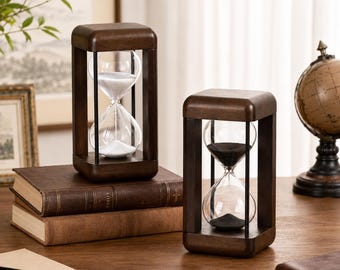 Puede incluir: Dos relojes de arena rectangulares con marcos de madera marrón oscuro y soportes de metal negro fino. Uno contiene arena blanca y el otro arena negra. Están colocados sobre una pila de dos libros marrones junto a un globo terráqueo.