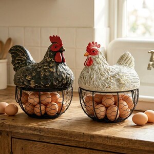 Puede incluir: Dos recipientes decorativos para huevos con forma de gallina. Uno es negro con cresta y barbilla rojas, el otro es blanco con detalles dorados y cresta y barbilla rojas. Cada uno tiene una cesta de alambre llena de huevos marrones.