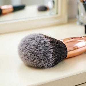 May include: A close-up of a makeup brush with a large, fluffy, gray and black bristle head. The handle is a shiny rose gold color. The brush is resting on a light-colored surface.