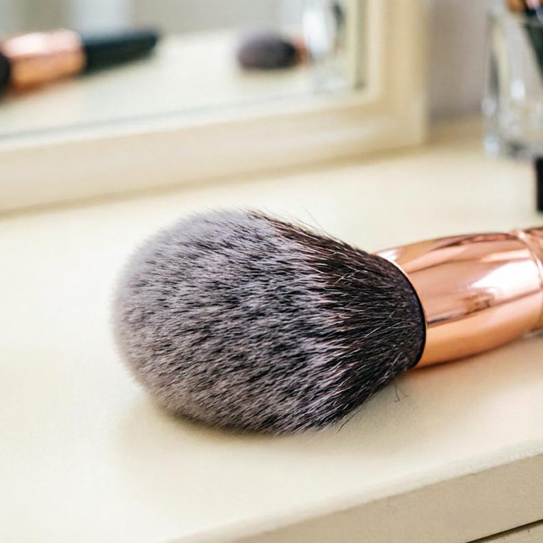 May include: A close-up of a makeup brush with a large, fluffy, gray and black bristle head. The handle is a shiny rose gold color. The brush is resting on a light-colored surface.