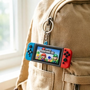 以下が含まれることがあります： Super Mario Bros. Wiiのロゴとキャラクターが表示された画面を持つ、ミニチュアゲーム機型のキーホルダー。青い左コントローラーと赤い右コントローラーを備え、銀色の金属製リングとチェーンに取り付けられています。