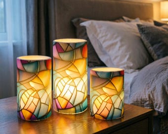 Puede incluir: Juego de tres velas LED decorativas con diseño de vidriera. Las velas presentan un colorido patrón de mosaico en tonos amarillos, naranjas, rosas y azules. Son cilíndricas y están sobre una mesita de noche de madera.