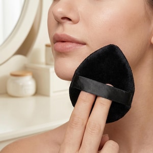 May include: A black, teardrop-shaped makeup sponge with a black strap is held near a person's face. The sponge has a soft, velvety texture. The background includes a white vanity and a jar with a wooden lid.